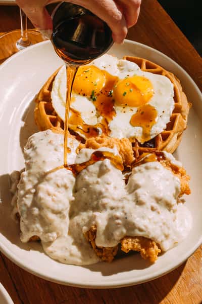 Southern-style brunch plate of crispy chicken with white gravy, waffles, and sunny-side-up eggs, being drizzled with maple syrup in a cozy restaurant setting