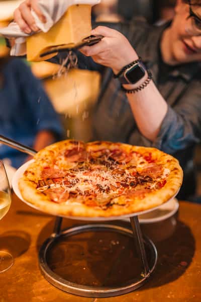 Close-up of a hot artisan pizza topped with cured meats and fresh-grated Parmesan being shaved tableside, capturing an interactive and upscale dining experience.