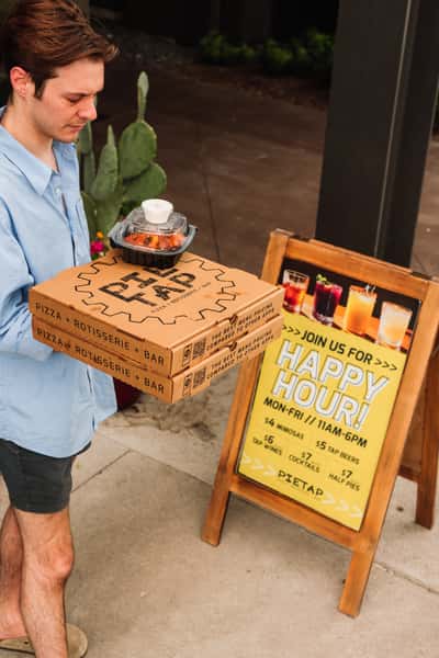 Person carrying pizza boxes and a side dish in front of a happy hour sign at Pie Tap Pizza Workshop + Bar, highlighting takeout options and drink specials like $4 mimosas and $5 craft beers.