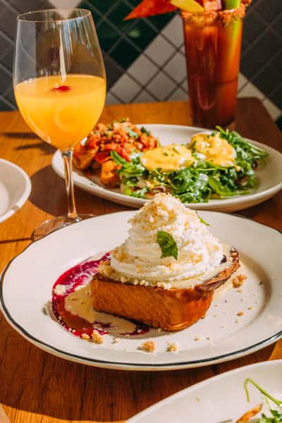 Dessert-style French toast topped with whipped cream and a mint leaf, drizzled with berry compote and cream sauce, surrounded by brunch drinks.