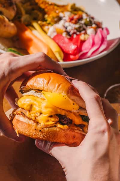 Close-up of a juicy breakfast burger with melted cheddar, fried egg, and crispy bacon held in two hands, with a colorful Mediterranean-inspired plate in the background.
