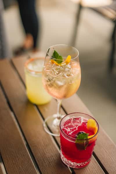 Trio of colorful cocktails on a wooden patio table, including a sparkling rosé spritz with orange twist, a lemon margarita with a salted rim, and a vibrant red berry drink garnished with mint and fruit.