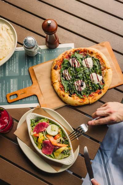 Flat-lay of an arugula and burrata pizza with a balsamic reduction, a colorful mixed salad, and a red berry cocktail on a wooden outdoor dining table.