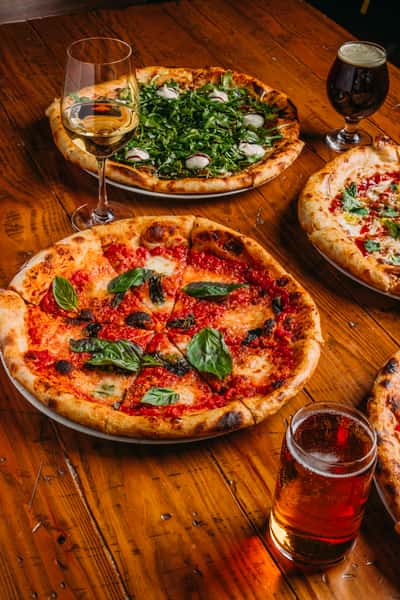 Four artisan pizzas topped with fresh ingredients like basil and arugula, paired with craft beer and white wine on a rustic wooden table.