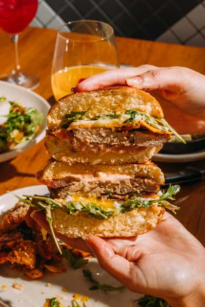 Close-up of a juicy chicken sandwich stacked with arugula, melted cheese, crispy bacon, and creamy sauce on a toasted bun, served with cocktails and salad at a modern eatery.