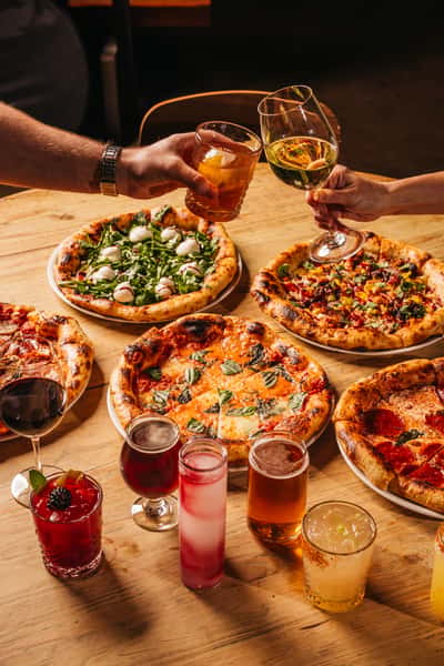 Image of several pizza and cocktails. There are two arms cheering with cocktails.