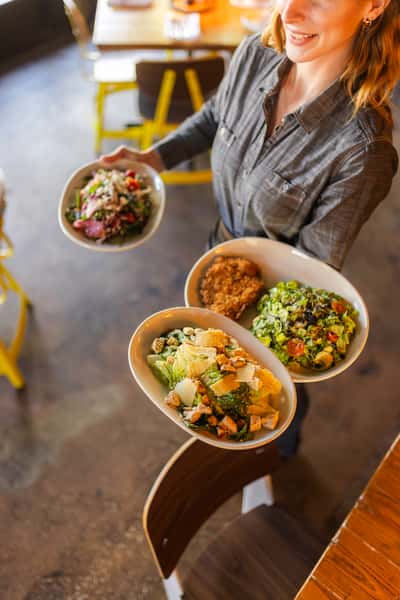 Smiling server carrying a trio of vibrant salads including Caesar, chopped, and a goat cheese and beet salad, presented in ceramic bowls at a bright, casual restaurant.