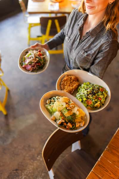 Server carrying a colorful assortment of fresh salads, including Caesar with grilled chicken, chopped greens with cherry tomatoes, and a quinoa bowl, highlighting healthy lunch options in a casual modern restaurant setting.