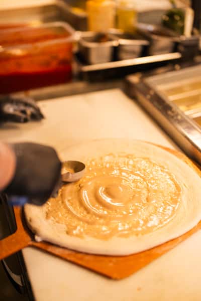 Close-up of pizza dough being spread with creamy white sauce on a wooden paddle in a restaurant kitchen.