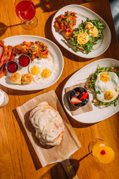 Brunch spread with eggs Benedict, sunny-side-up eggs with biscuits and bacon, mixed greens, a large iced cinnamon roll, fresh berries, and colorful brunch cocktails.