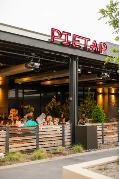 Outdoor patio dining at Pie Tap restaurant, with groups of guests enjoying meals beneath a covered terrace and illuminated red neon signage.
