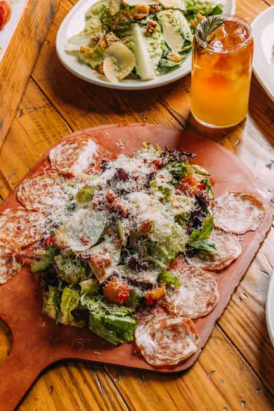 Close-up of an Italian salumi salad topped with grated Parmesan and surrounded by thin slices of salami, served with a rosemary cocktail and Caesar salad in the background.