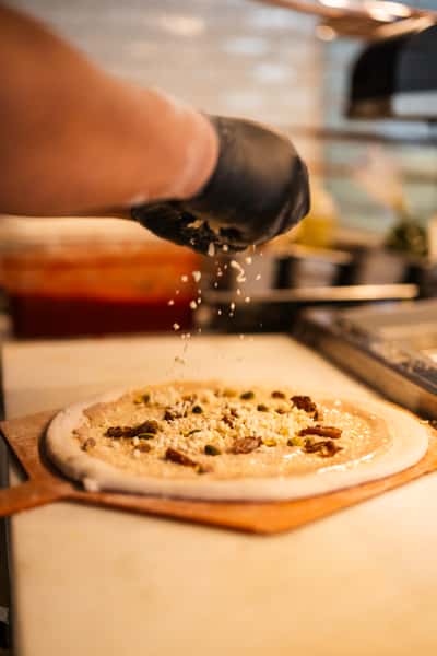 Close-up of pizza preparation with cheese and toppings being sprinkled over white sauce on hand-stretched dough in a professional kitchen.