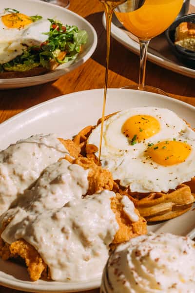 Fried chicken topped with white gravy, waffles with fried eggs, and syrup being poured over them.