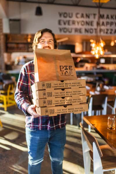 Smiling customer holding stacked takeout boxes and a brown bag from Pie Tap Pizza Workshop + Bar, inside a cozy pizzeria with a humorous wall quote about pizza and happiness.