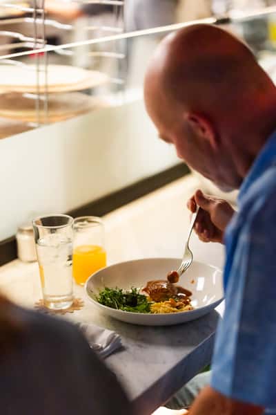 Man dining solo at a modern restaurant, enjoying a plated entrée with greens and pasta, seated at a marble counter with water and juice
