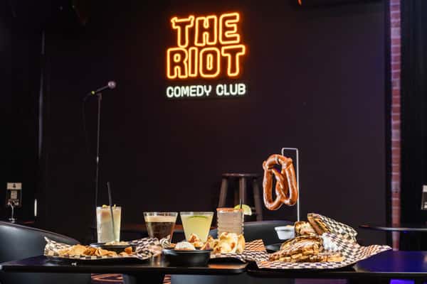 Multiple dishes and drinks in front of a neon sign for The Riot
