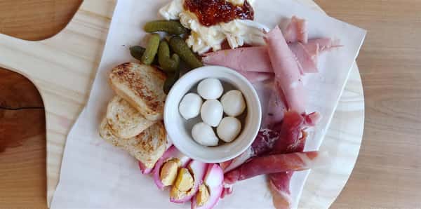A charcuterie board with prosciutto, mortadella, fresh mozzarella, brie, fig jam, pickled brussels sprouts, pickled egg, cornichons, and roasted bread