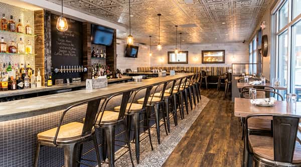 Modern restaurant interior with a bar counter, metal stools, and dining tables, textured ceiling, and wood accents
