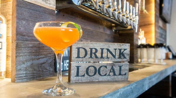 Orange cocktail with a lime twist on a concrete bar, in front of beer taps and a "Drink Local" sign