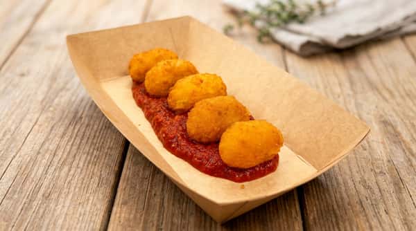 Five golden-fried croquettes served in a line on a bed of red marinara sauce in a disposable paper tray.