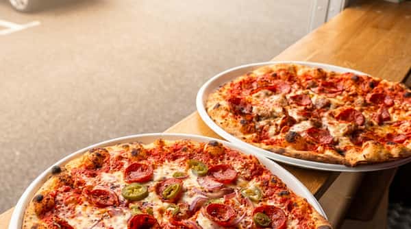 Two pizzas on white plates on a wooden counter with a street view: one with pepperoni and jalapenos, the other with pepperoni and sausage.