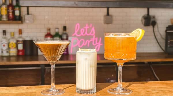 Three cocktails on a wooden bar counter with a pink "let's party" neon sign and white brick background.