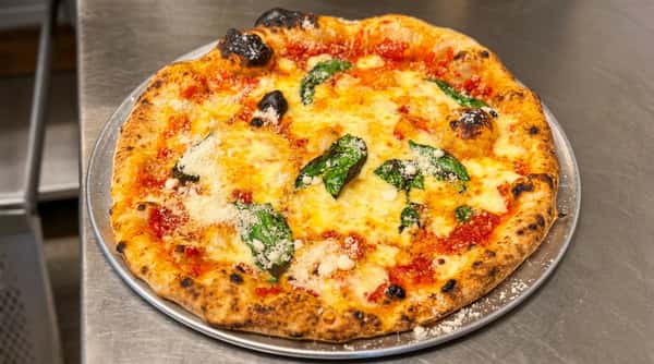 Neapolitan pizza with charred crust, melted mozzarella, red sauce, basil leaves, and grated cheese on a metal tray.
