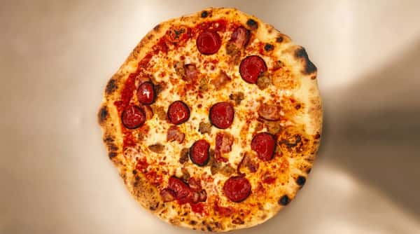 Pepperoni, sausage, and bacon pizza with melted cheese and a bubbly crust on a light background.