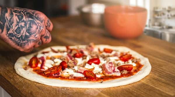 Uncooked pizza with cherry tomatoes, mozzarella, bacon, sausage, and pepperoni, prepared by a tattooed hand.