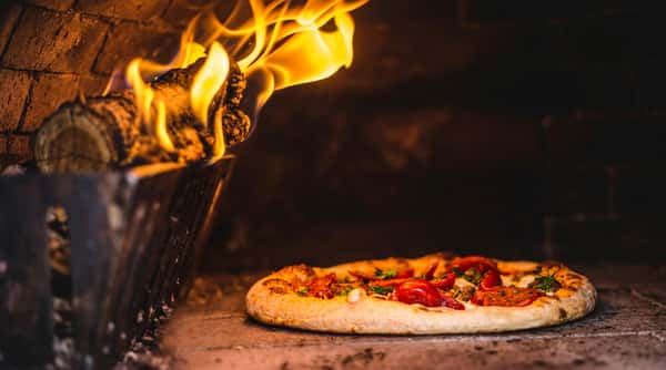 Pizza baking in a dark, wood-fired oven with visible flames on the left.