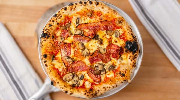 Neapolitan pizza with crispy bacon strips, sliced mushrooms, melted mozzarella, and charred crust on a metal tray.