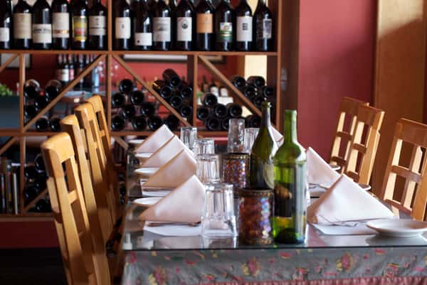A cozy, warmly lit dining table set with crisp white napkins, glassware, and rustic accents, framed by walls of neatly displayed wine bottles for an inviting Italian atmosphere.