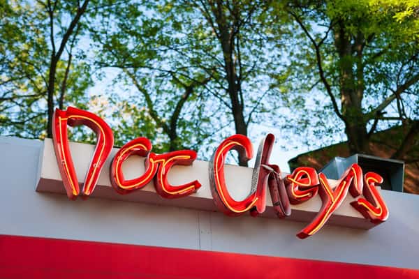 Red neon sign reading "Doc Chey's" mounted on a white and red building exterior, with green trees in the background.