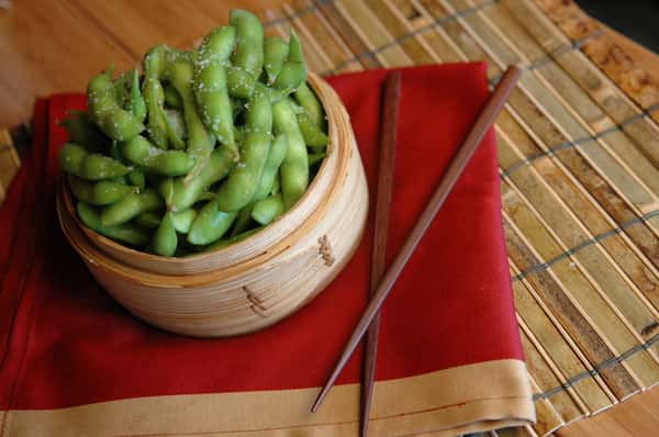Steamed edamame sprinkled with sea salt served in a bamboo bowl on a red napkin with wooden chopsticks and a bamboo mat.