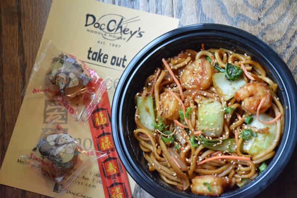Shrimp lo mein with bok choy and sesame seeds in a black takeout bowl, with fortune cookies and Doc Chey's takeout menu nearby.