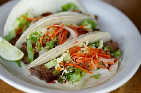 Three soft tacos filled with grilled beef, shredded lettuce, pickled carrots, onions, and a drizzle of hot sauce, served with lime.