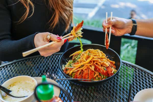 Two people using chopsticks to share a bowl of crispy noodle salad with sliced meat at an outdoor restaurant table.