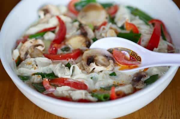 Bowl of Thai coconut soup with mushrooms, red bell peppers, chicken, green onions, and a ceramic spoon on the side.