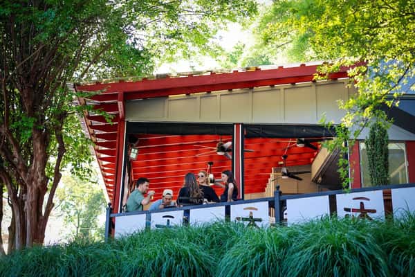 Group of people dining on a shaded outdoor patio with a red ceiling, surrounded by lush green trees and tall grass.