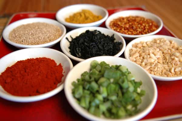 A colorful assortment of toppings and spices in small white bowls, including scallions, peanuts, seaweed, sesame, and chili flakes.