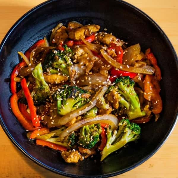 Stir-fried chicken with broccoli, red bell peppers, onions, and sesame seeds in a savory sauce, served in a black bowl.