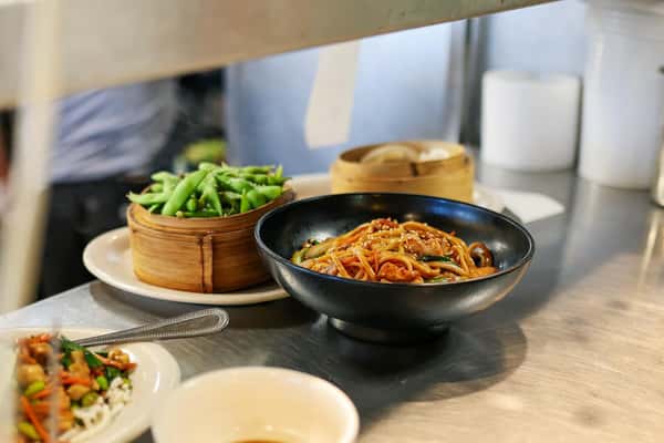 Steaming edamame, lo mein noodles with vegetables, and dumplings plated and ready for service at a restaurant kitchen pass.