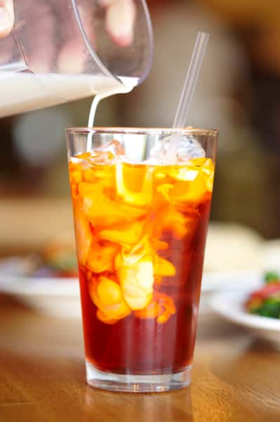 Cream being poured into a glass of iced Thai tea, creating a swirling effect, with a straw and blurred food dishes in the background.