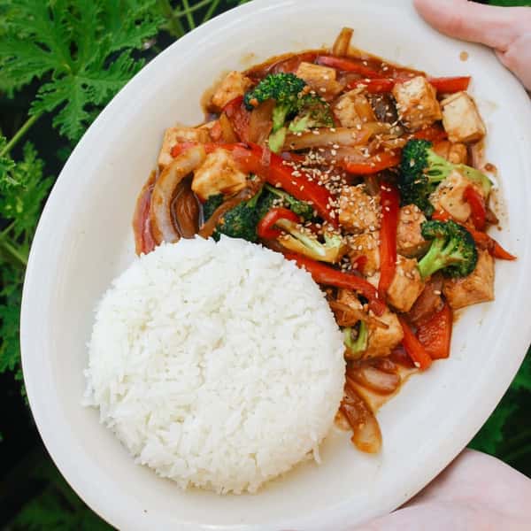 White rice served with stir-fried tofu, broccoli, red bell peppers, and onions topped with sesame seeds on a white oval plate.