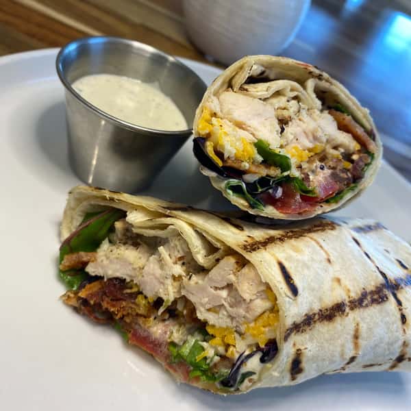 Grilled chicken wrap filled with tender chicken, cheddar cheese, bacon, fresh greens, and tomatoes, served with a side of creamy ranch dressing on a white plate.