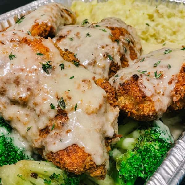 A serving of crispy fried chicken topped with creamy gravy, served alongside mashed potatoes and steamed broccoli, garnished with fresh thyme.