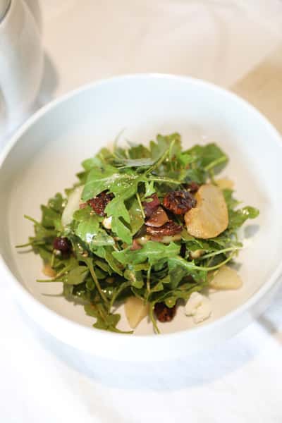 Fresh arugula salad with dried tomatoes, sliced almonds, and crumbled feta, drizzled with a light vinaigrette dressing, served in a white bowl