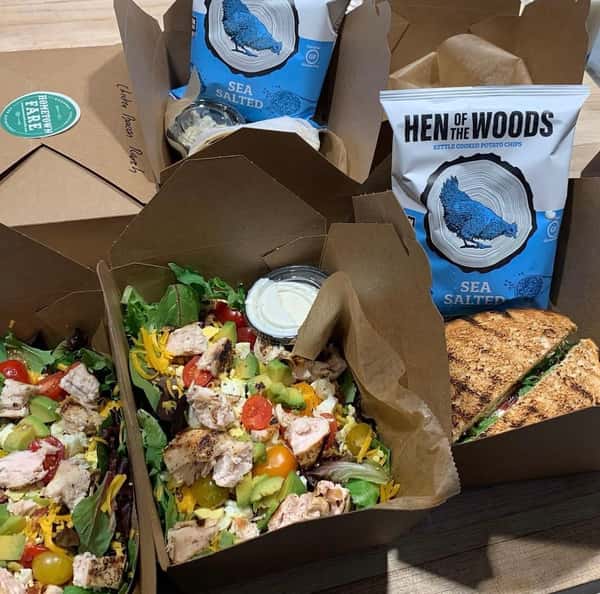 A lunch spread featuring two fresh salads with grilled chicken, avocado, tomatoes, and shredded cheese, accompanied by sea salted kettle chips and a sandwich.