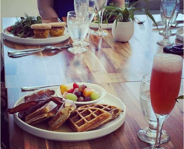 A plate of crispy bacon, waffles, and fresh fruit salad with a sparkling beverage in a cozy restaurant setting.
